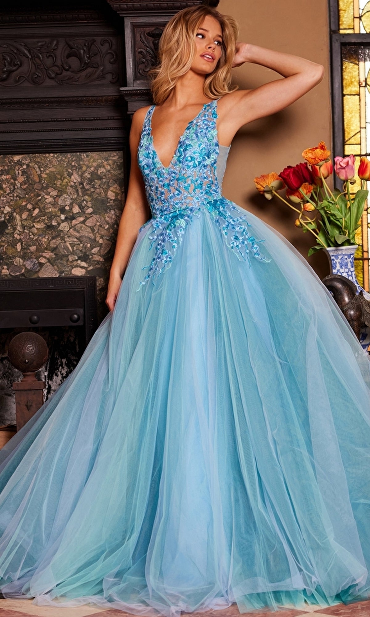 A woman in a sleeveless, blue Jovani Tulle Prom Ball Gown with floral embroidery stands indoors by a fireplace and a vase of vibrant tulips, her dress radiating elegance.